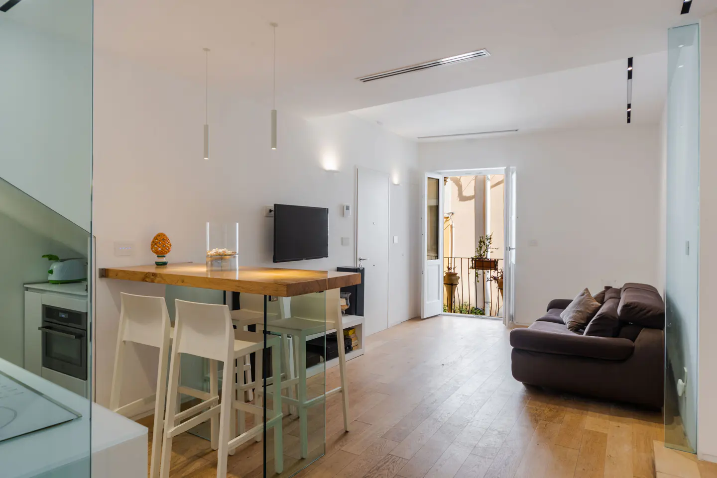 Bright, modern apartment interior with a wooden breakfast bar, white stools, and a brown sofa near an open balcony door.