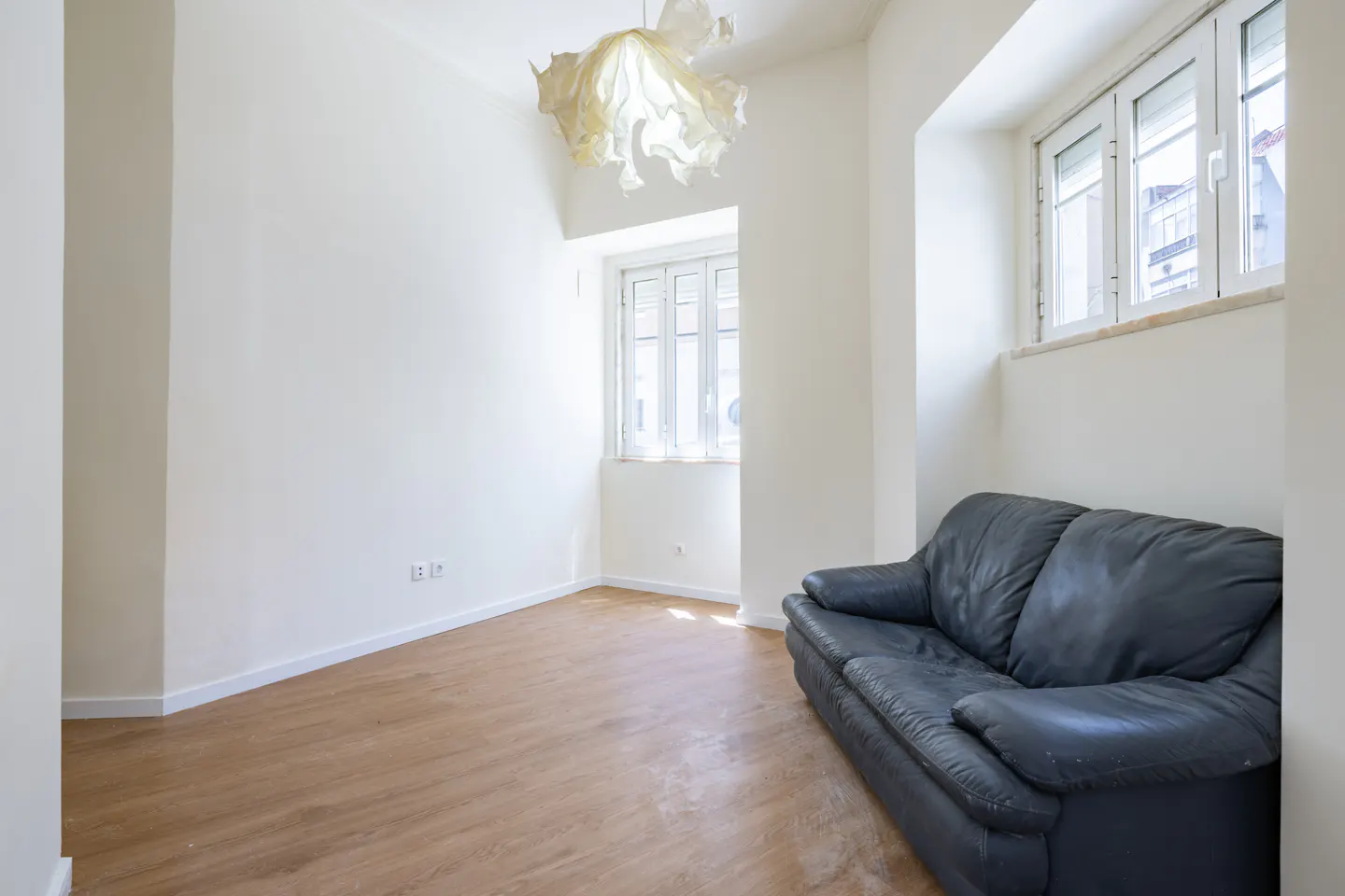Bright, empty room with wood floors, white walls, and a black leather couch. A modern light fixture hangs from the ceiling. Two windows let in natural light.