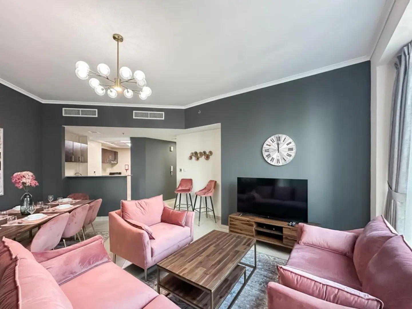 A modern living room with pink velvet sofas, a wooden coffee table, and a TV. A dining area with pink chairs is visible in the background.