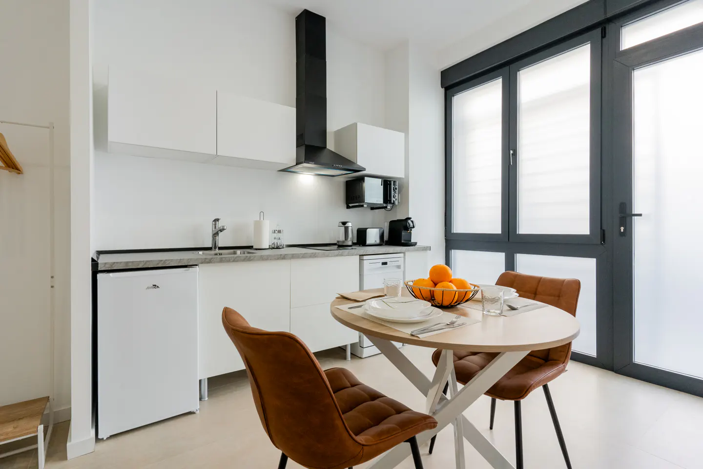 Modern kitchen with white cabinets, black range hood, and a round dining table with two brown chairs and a bowl of oranges.