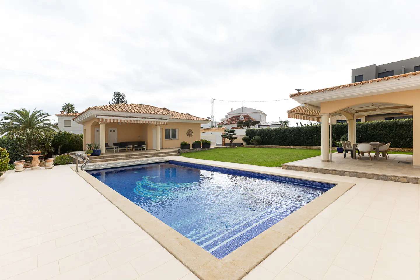 A backyard with a blue tiled pool, a beige house with a tiled roof, and a covered patio with a table and chairs.