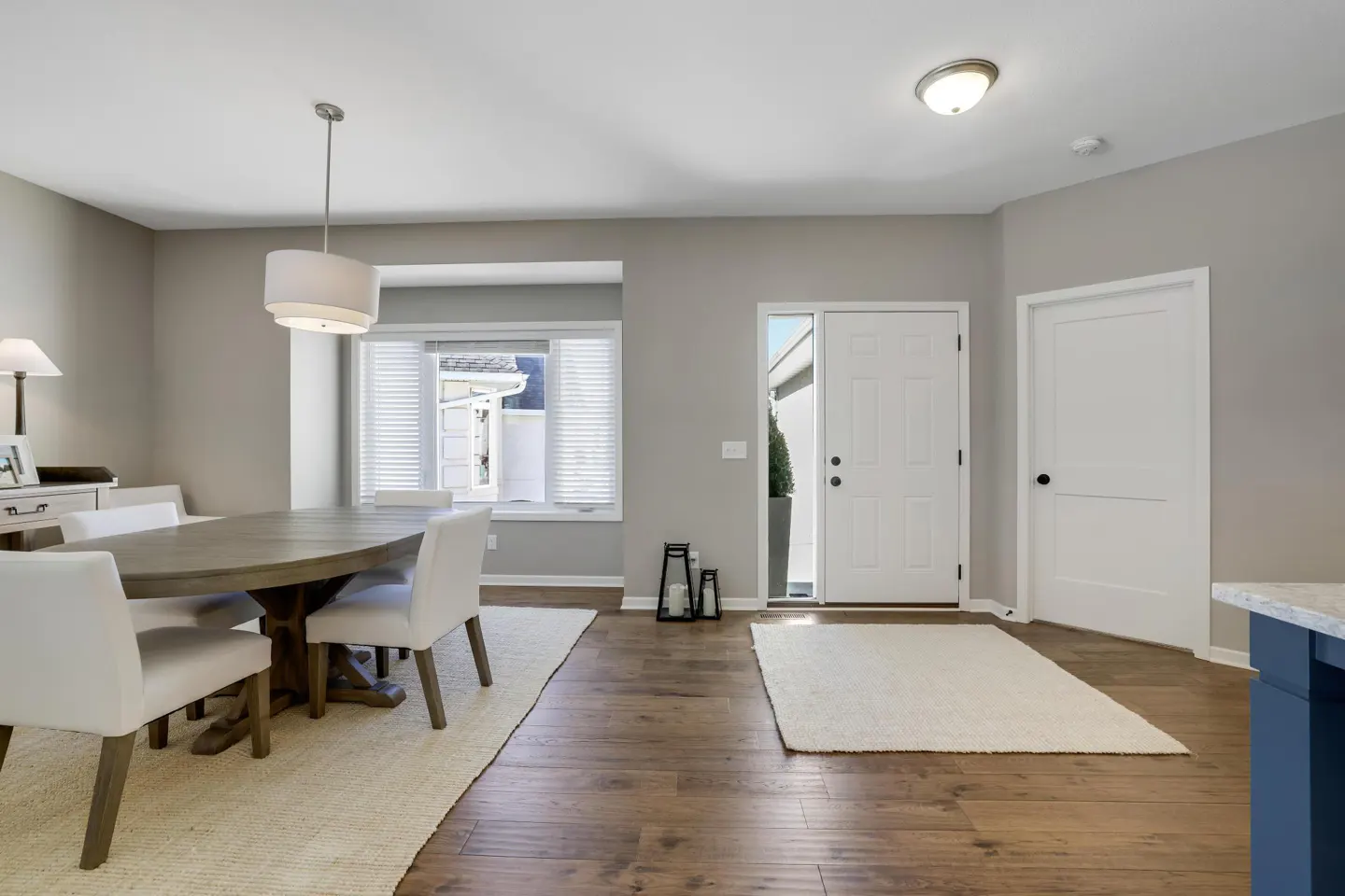 Bright, neutral entryway with wood floors, white door, and round wood table with white chairs. A window with blinds is visible.