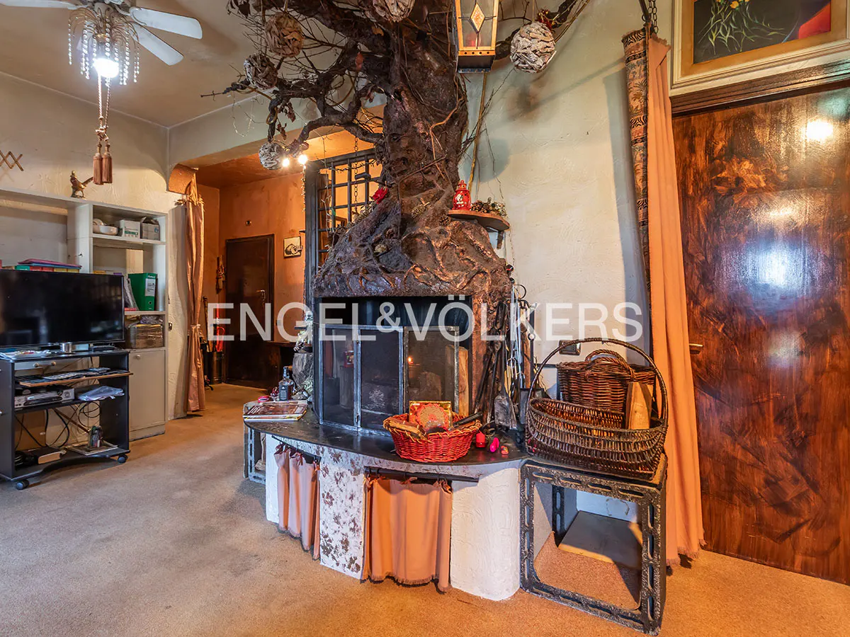 Living room with a tree-like fireplace, TV stand, and a ceiling fan with lights. The walls are beige and there is a wooden door on the right.