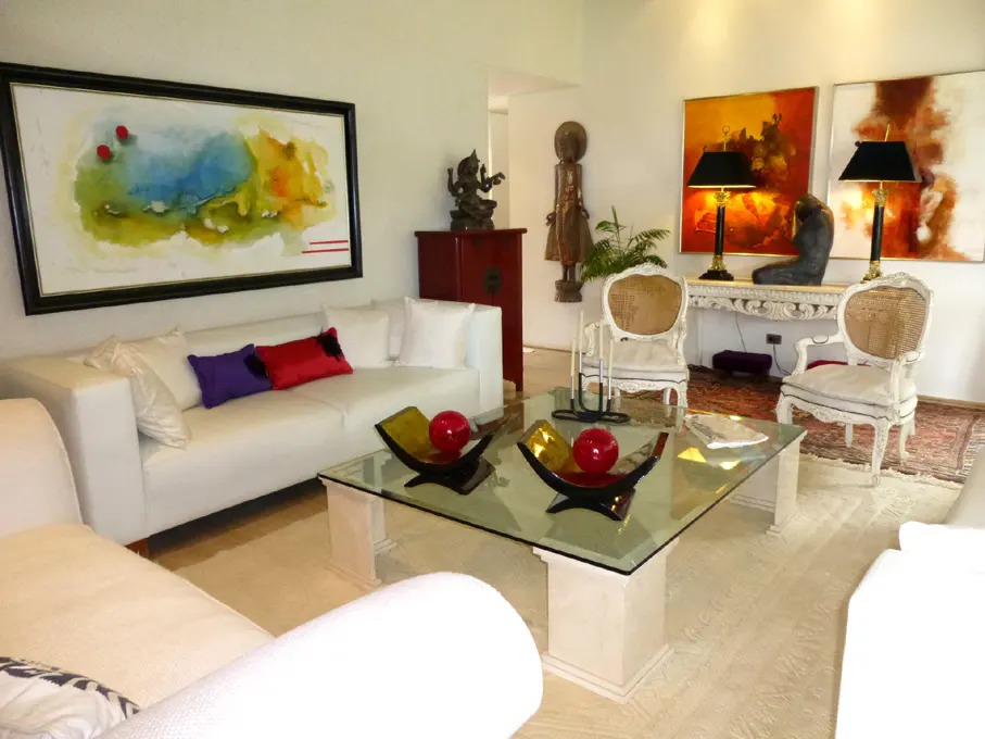 Bright living room with white sofas, colorful art, and a glass coffee table with decorative bowls. Statues and lamps add to the elegant decor.