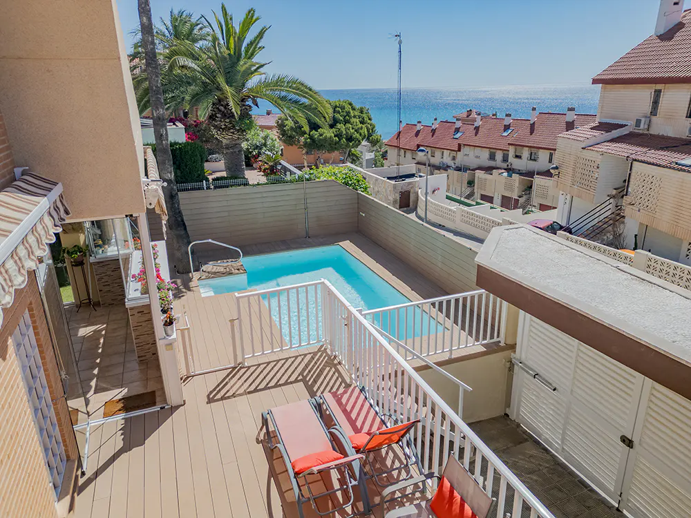 Aerial view of a pool with lounge chairs on a deck, palm trees, and the ocean in the background.