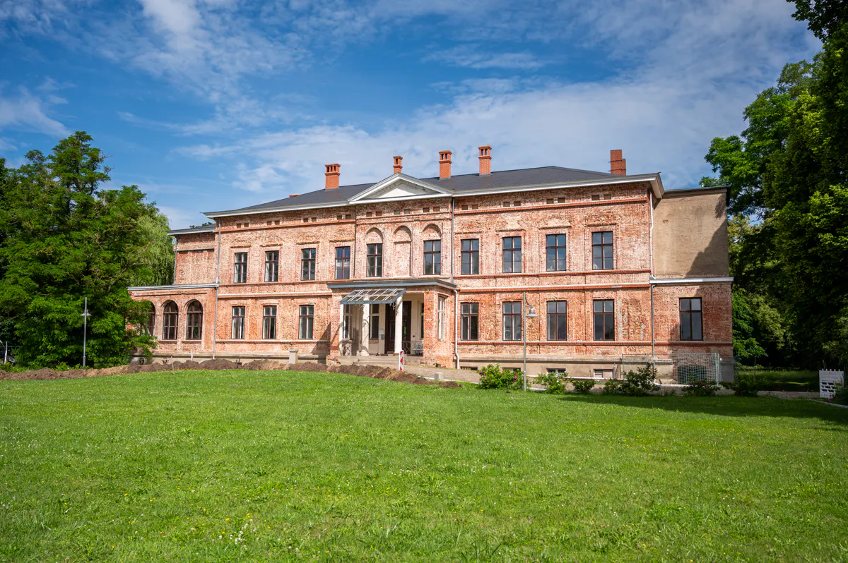 A three-story red brick building with a black roof and chimneys, surrounded by a green lawn and trees under a blue sky.