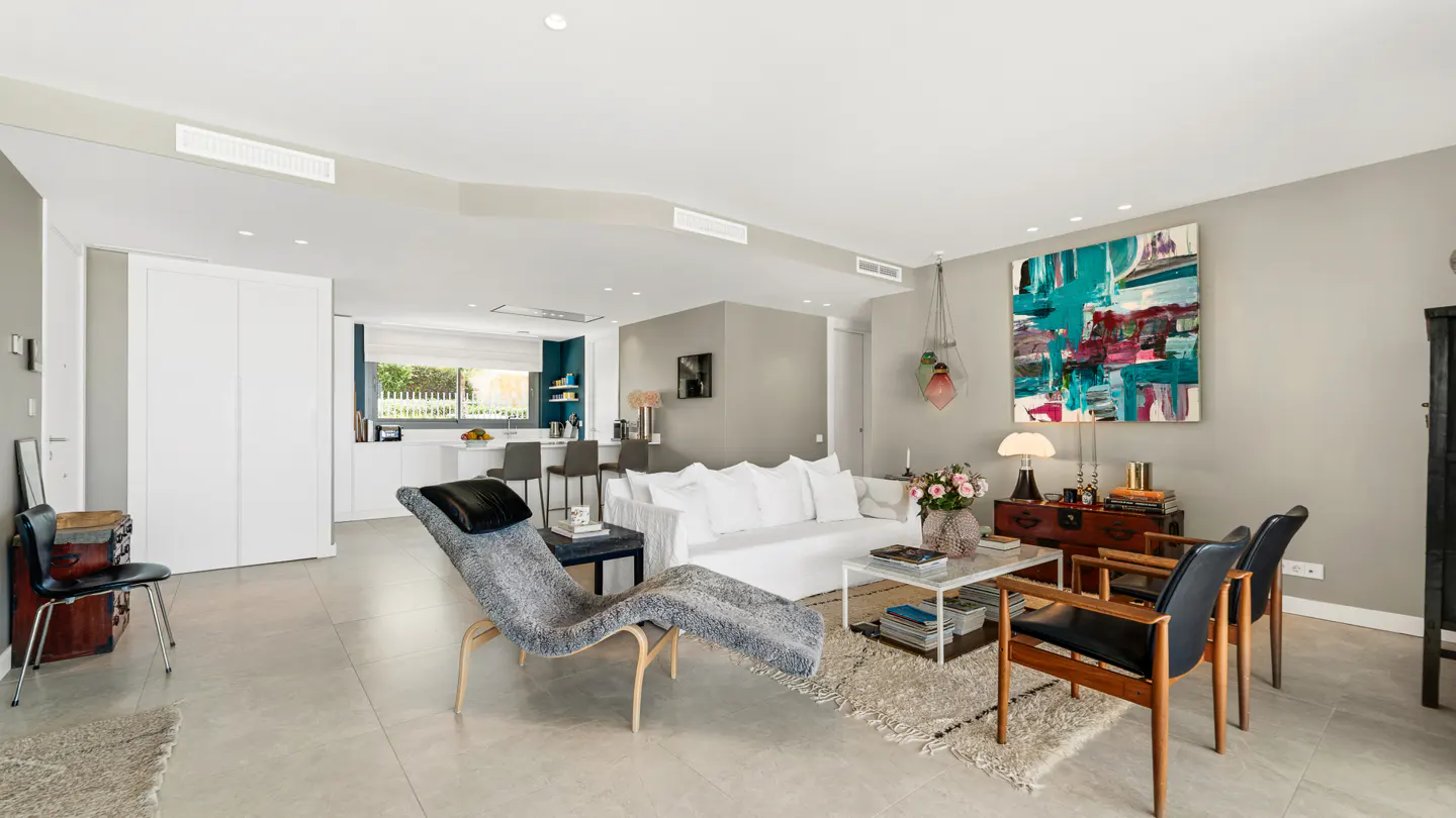 Open-concept living room with a white sofa, chaise lounge, and colorful abstract art. Kitchen visible in the background.