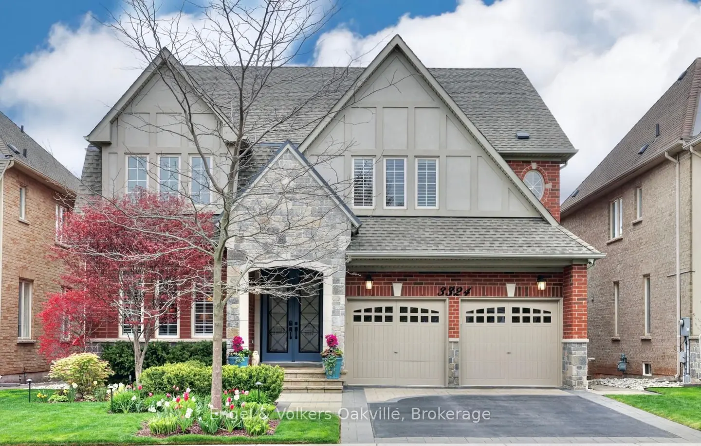 Two-story house with gray roof, brick and stone facade, and two-car garage. A tree and colorful flowers are in the front yard.