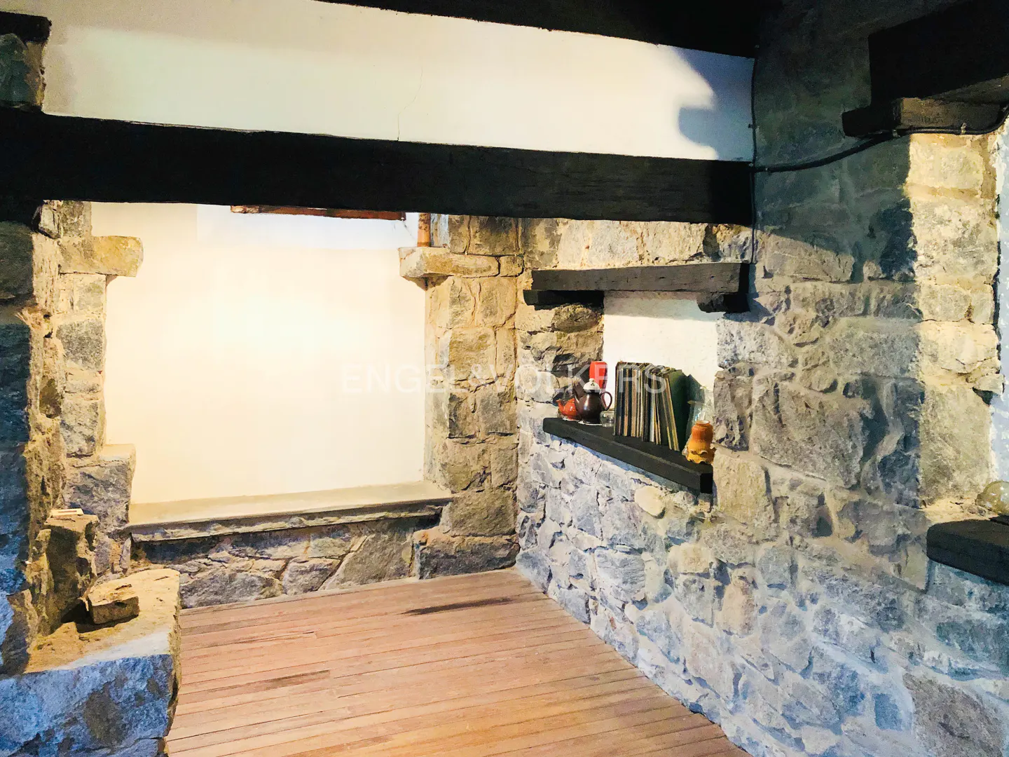 Stone wall interior with wooden floors, black beams, and a shelf with books and decorative items.