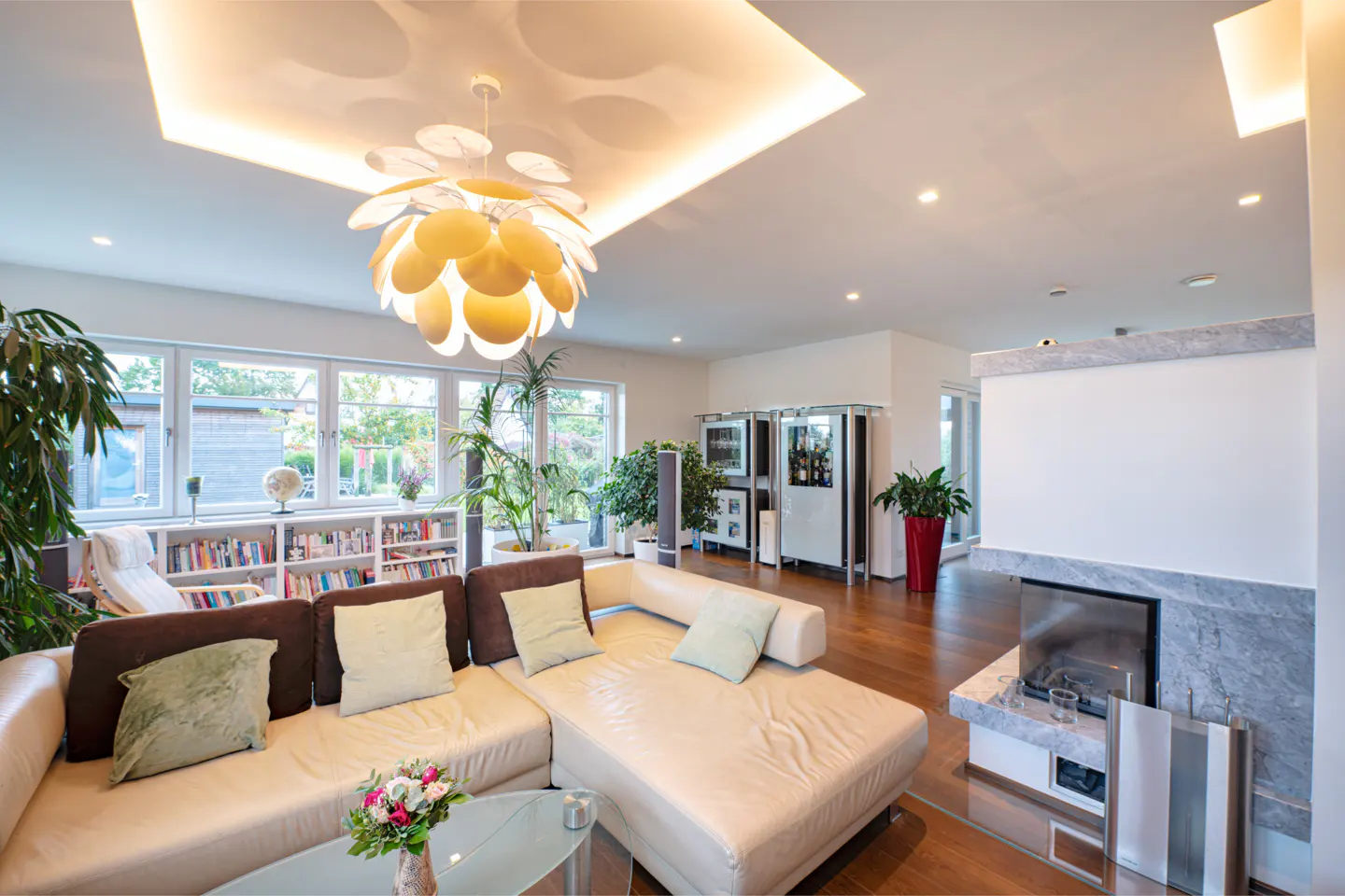 Bright living room with a large beige sectional sofa, a modern chandelier, and a fireplace with a marble surround. Bookshelves line the wall near the windows.