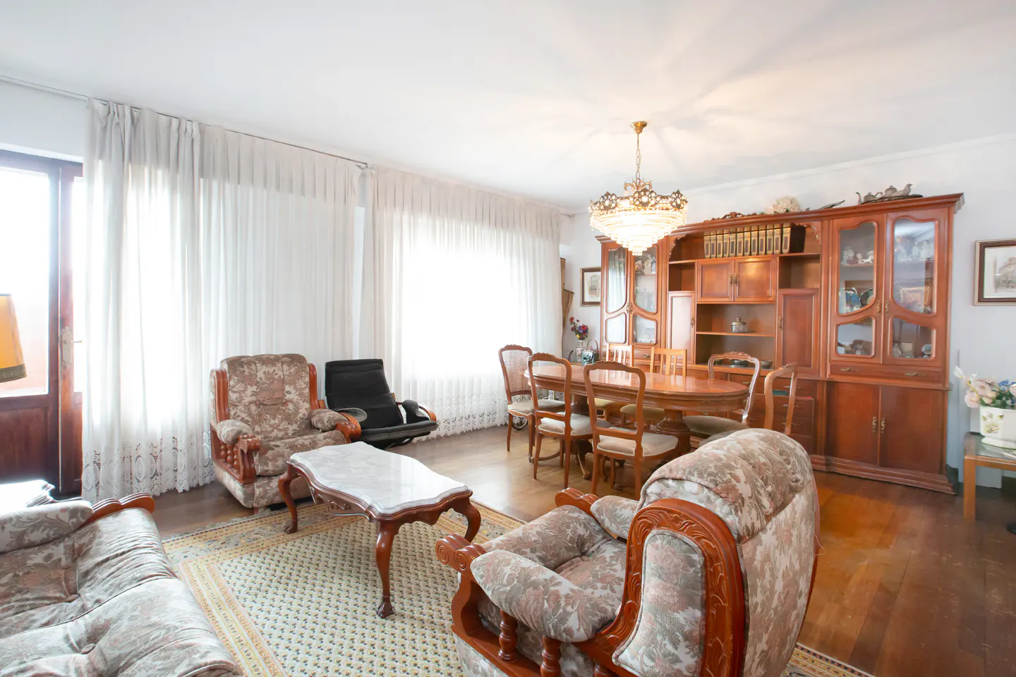 A bright living room with floral armchairs, a marble-top coffee table, and a wooden dining set under a chandelier.