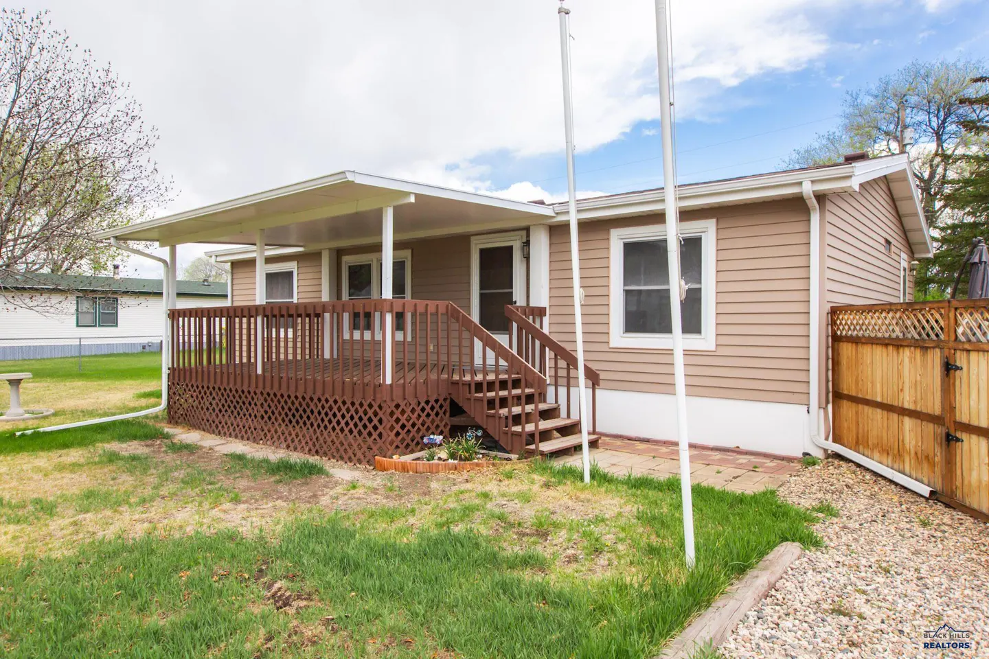 Tan-sided house with a brown deck and white awning. Green grass and a blue sky with clouds in the background.