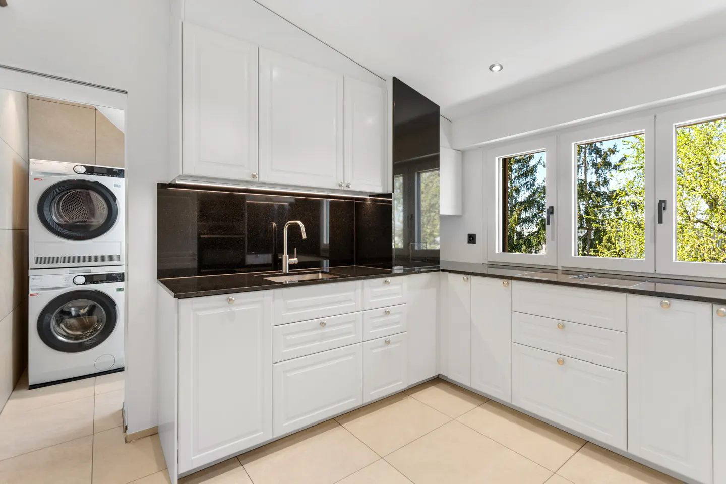 A modern kitchen with white cabinets, black countertops, and a stacked washer and dryer in the background. Three windows offer a view of green trees.
