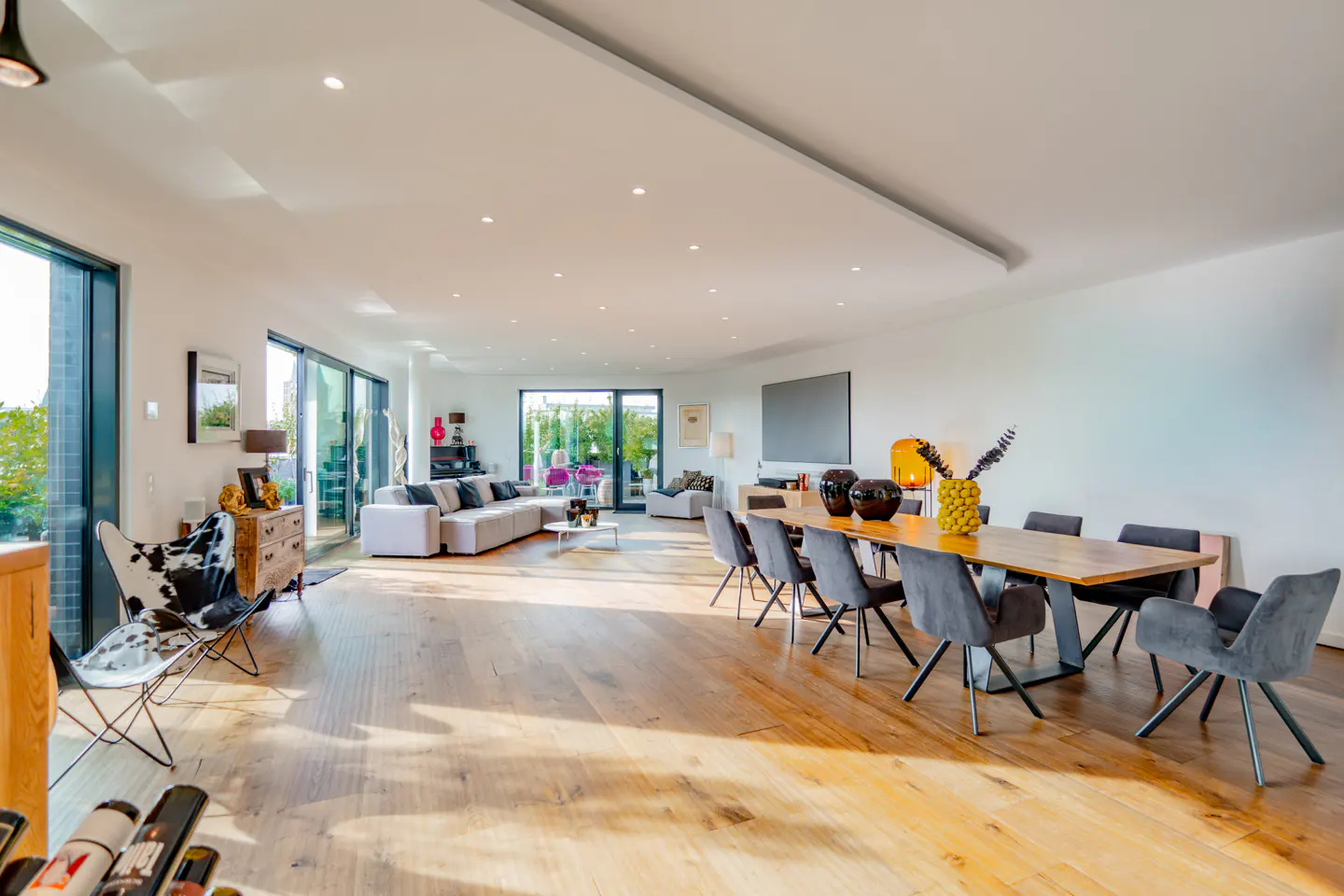 Bright, open-plan living and dining area with light wood floors. A long wood table with gray chairs sits near a white sofa and large windows.