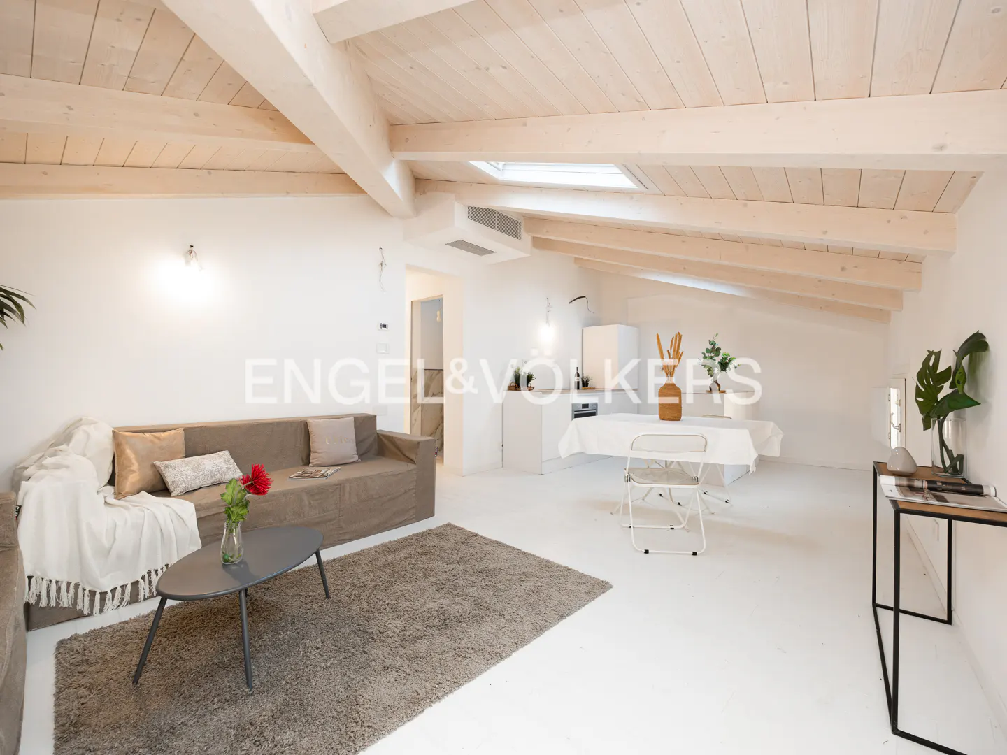 Bright attic living room with white walls, exposed wooden beams, a brown sofa, gray rug, and a dining area in the back.