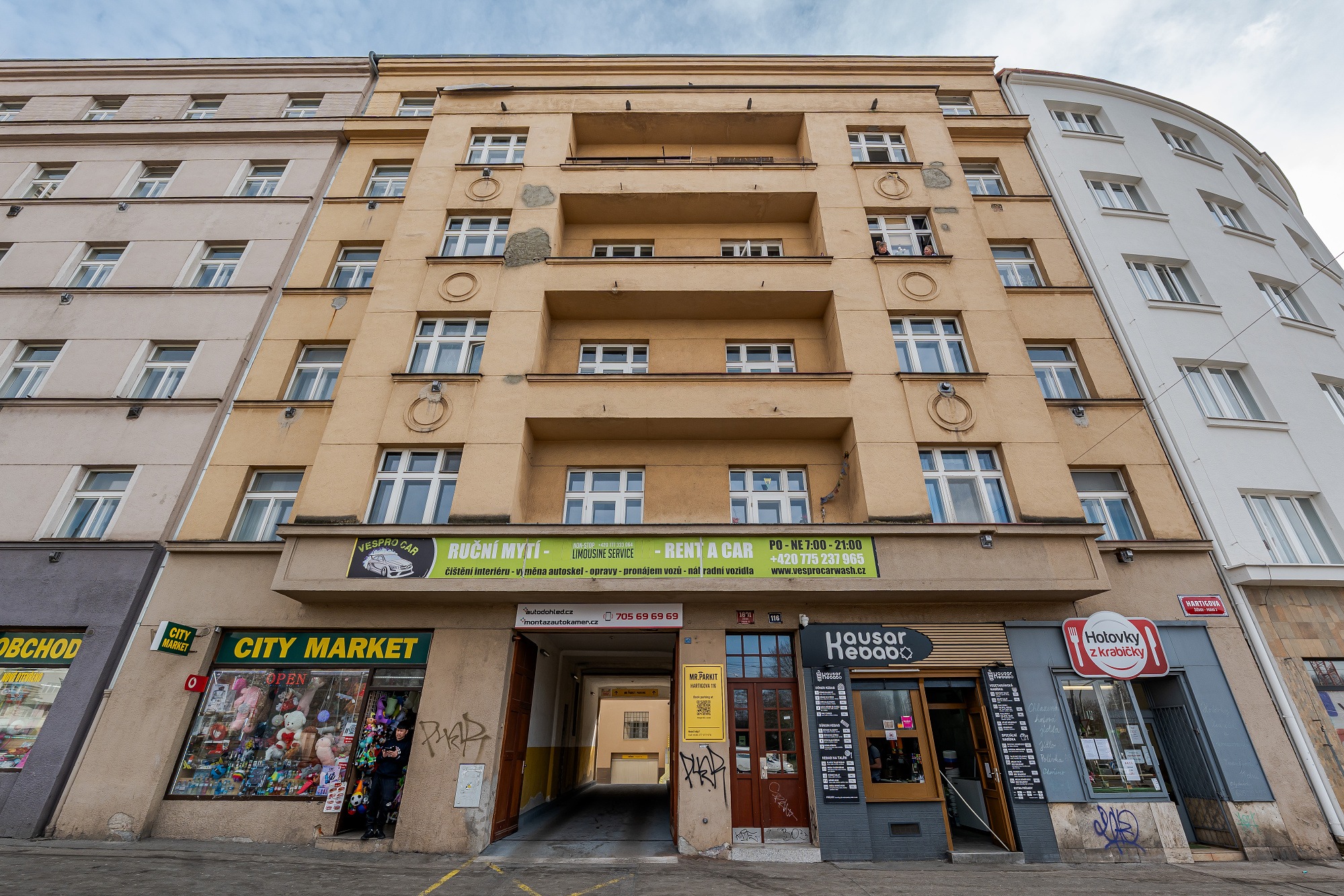 A tan multi-story apartment building with a ground-floor car wash, City Market, and kebab shop on a city street. [AI-generated text]
