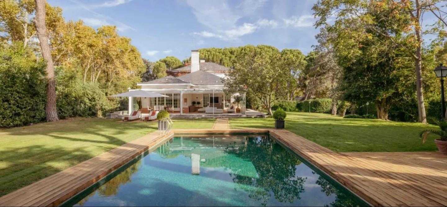 A backyard view of a white house with a pool, wooden deck, green lawn, and trees under a blue sky.