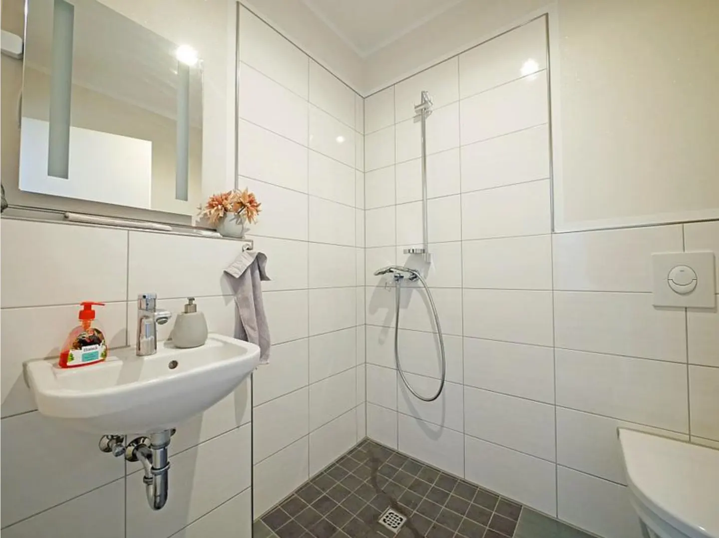 Bright bathroom with white tiled walls, gray tiled floor, sink, mirror, and shower.