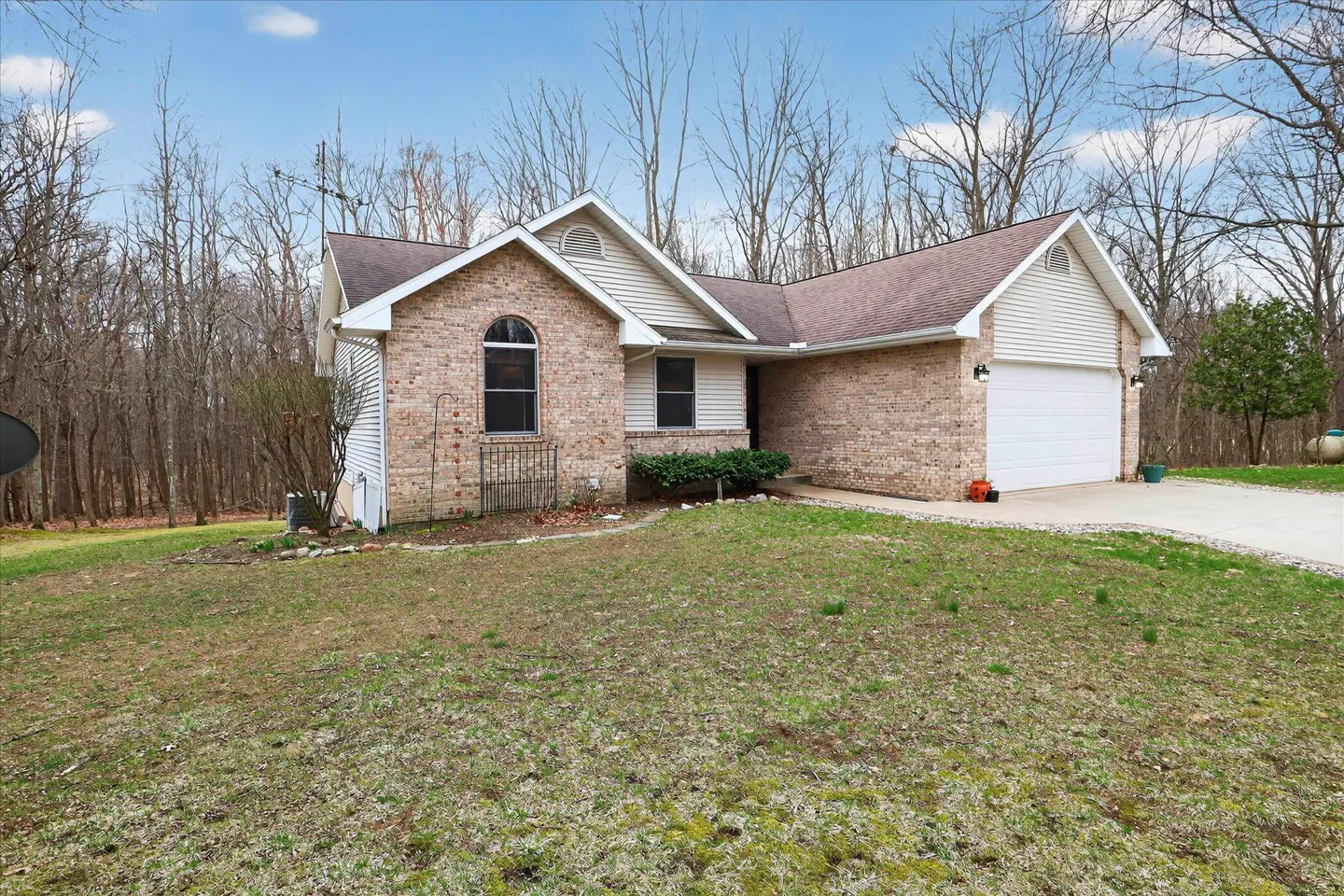 A single-story brick house with a white garage door and a lawn in front of a wooded area.