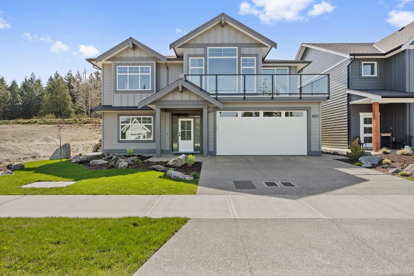 Two-story gray house with white trim, a balcony, and a white garage door. A green lawn and landscaping surround the house.