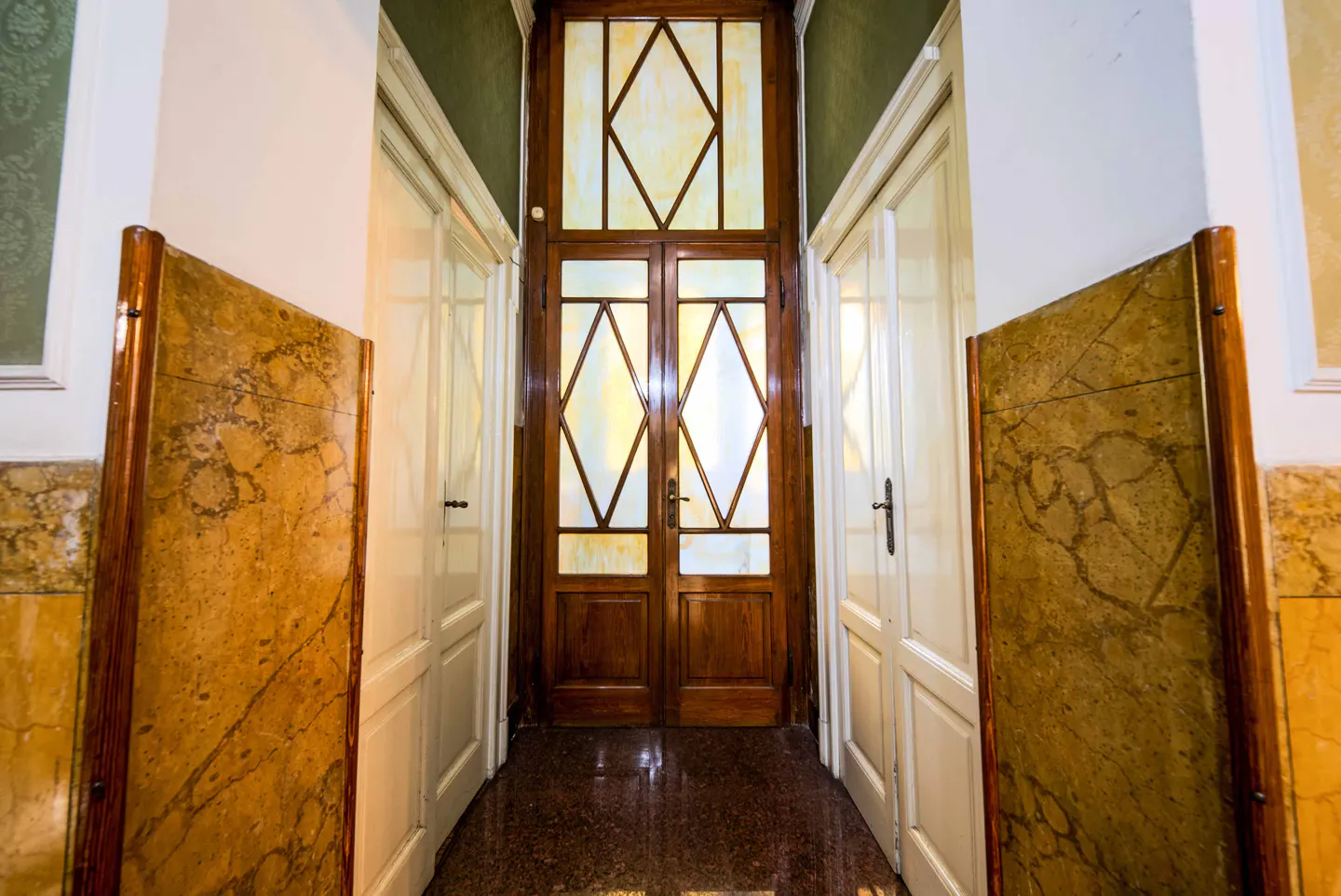 Hallway with marble floor, white walls, and a dark wood door with diamond-shaped glass panels at the end. White doors on either side.