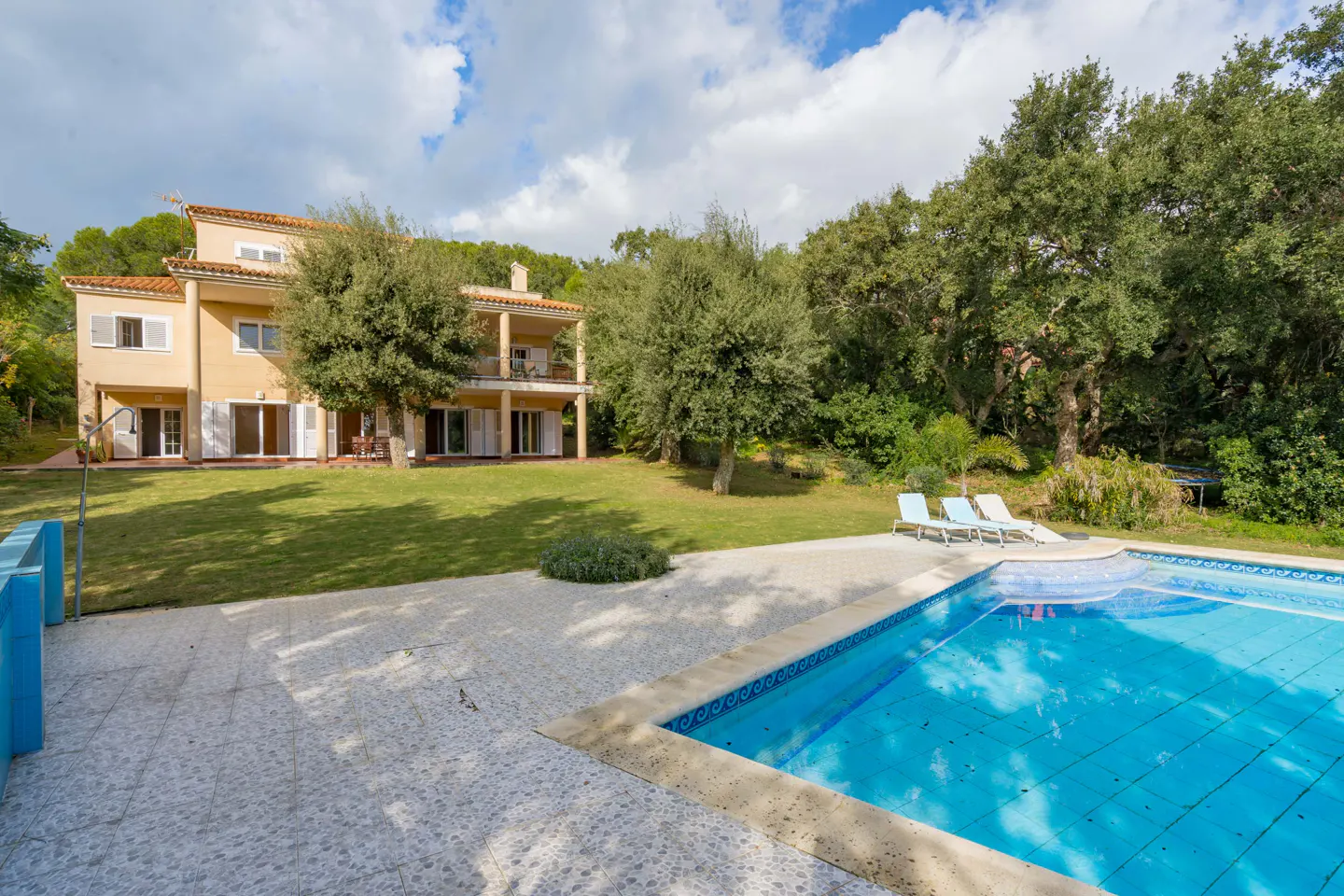 A tan two-story house overlooks a blue tiled pool and lawn with lounge chairs. Trees and a cloudy sky are in the background.
