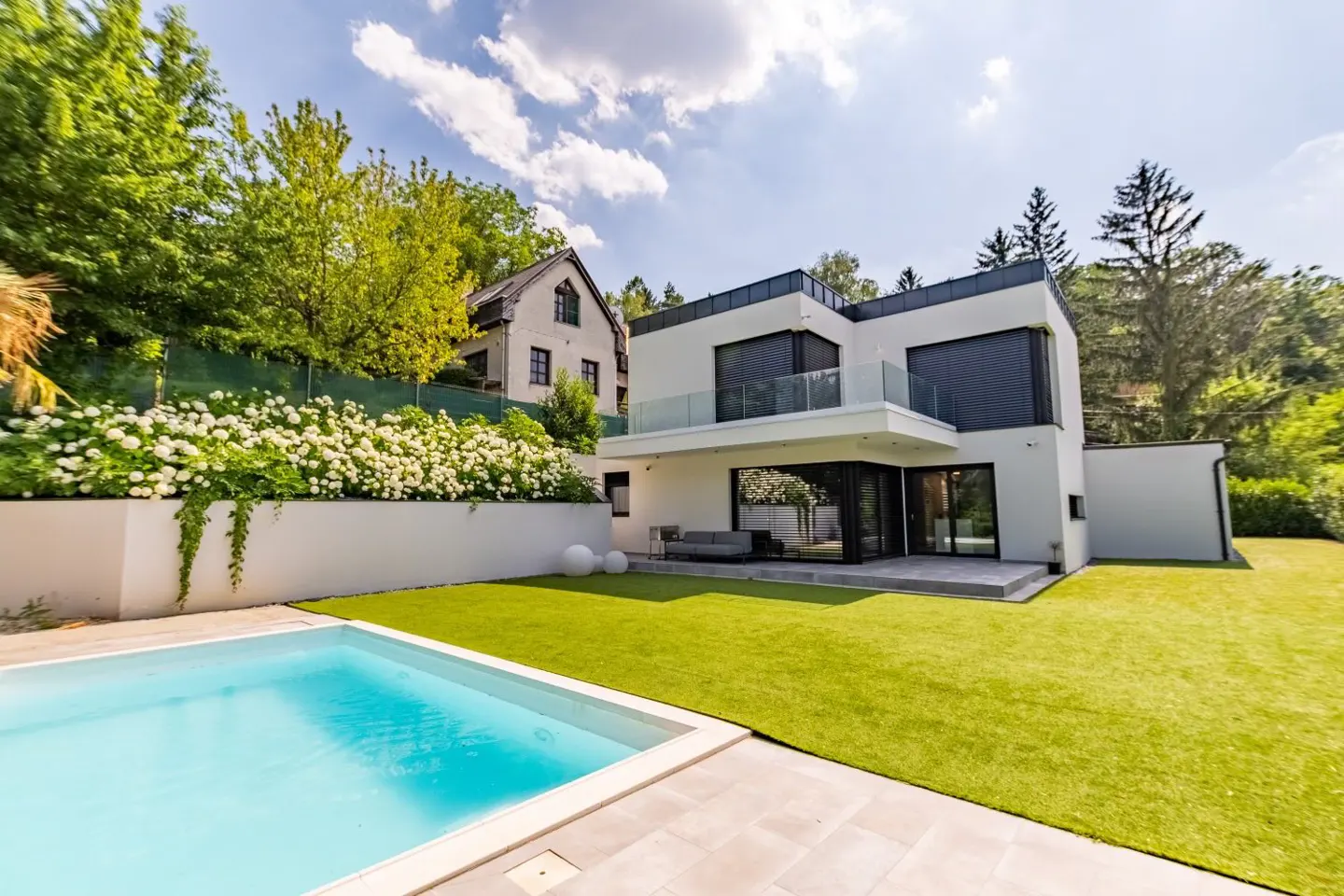 Modern white house with a pool and green lawn. A white wall with flowers separates the property from the neighbor's house.