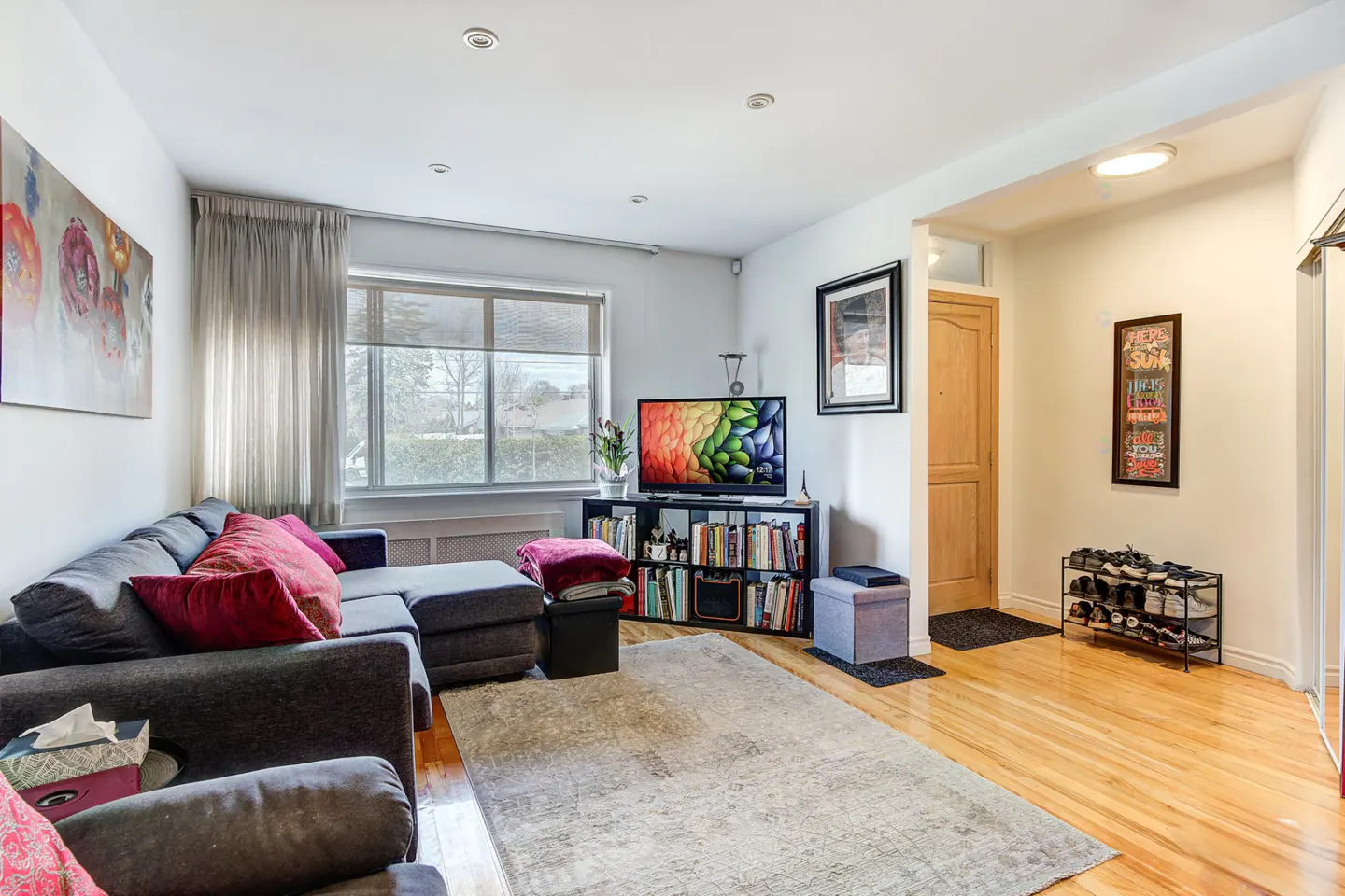Living room with a gray sectional sofa, pink pillows, a TV on a bookshelf, and a light gray rug on a wood floor.