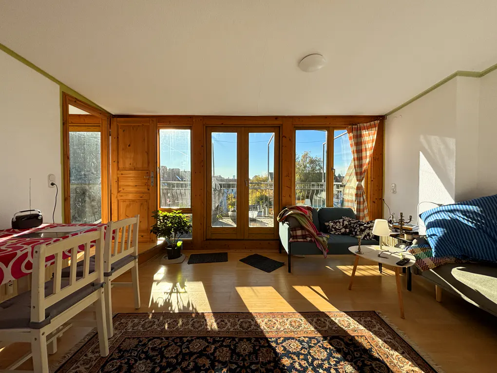 Sunlit living room with wooden doors to a balcony. A dining table sits to the left, and a blue sofa is on the right. A patterned rug covers the floor.