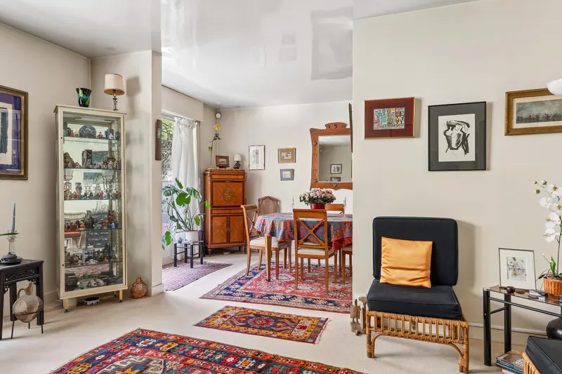 A living room with a display cabinet, dining table, and black chair. Colorful rugs cover the floor. Art hangs on the walls.