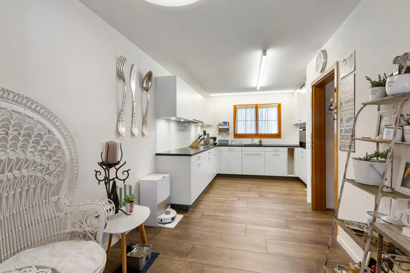Bright kitchen with white cabinets, wood floors, and decor. Silverware art hangs on the wall next to a white wicker chair.