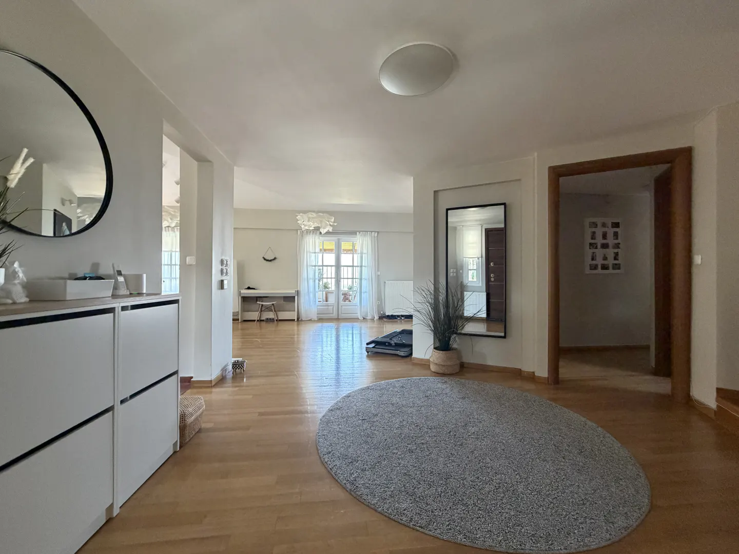 Bright, open-plan home interior with light wood floors, a large grey oval rug, white cabinets, and mirrors.