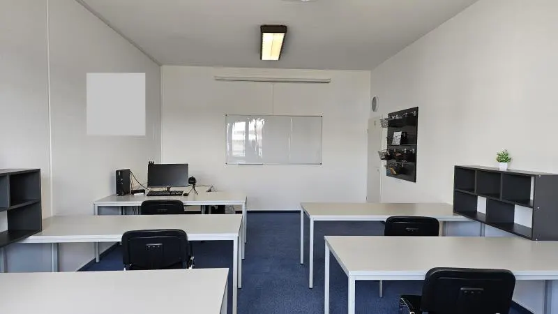 Bright office space with white walls, blue carpet, and white desks with black chairs. A whiteboard and computer are visible.