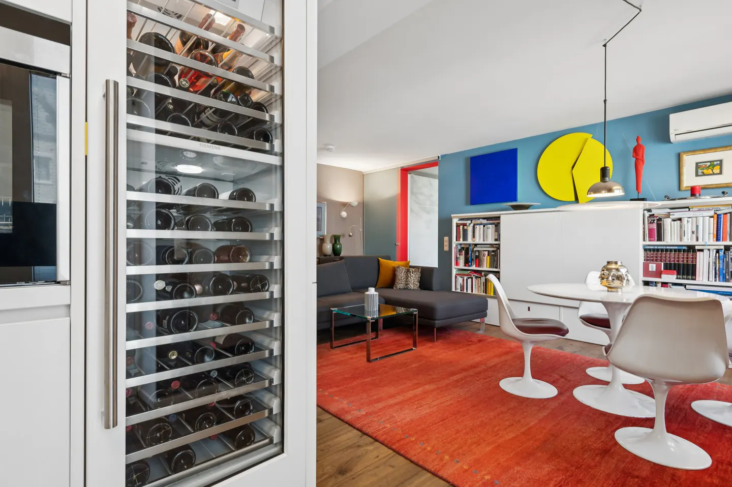 A modern living room with a wine fridge, gray sofa, orange rug, and a dining table with tulip chairs. The walls are blue with colorful art.
