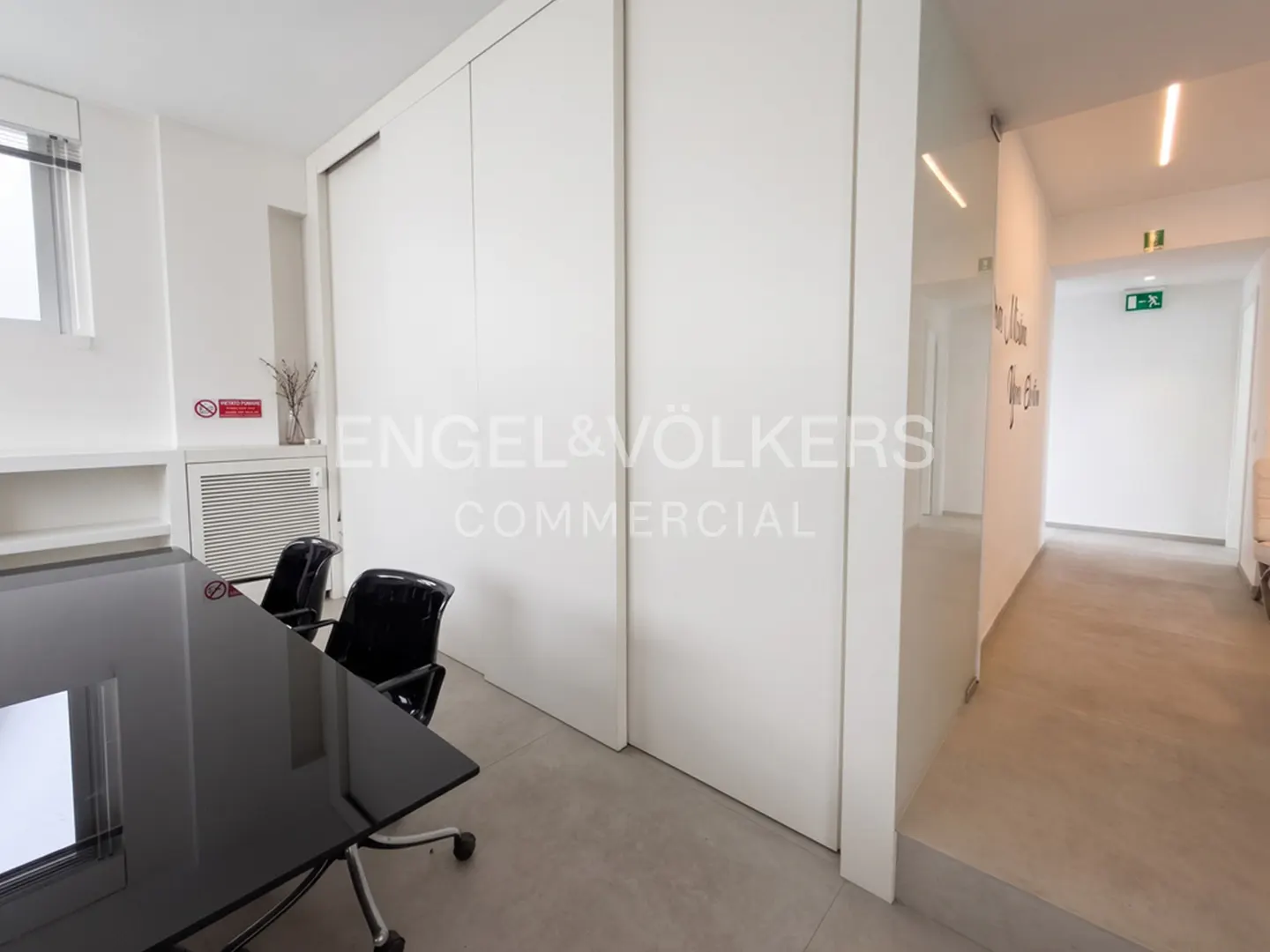 A bright office space with a black conference table, chairs, and white walls. A hallway is visible with an exit sign.