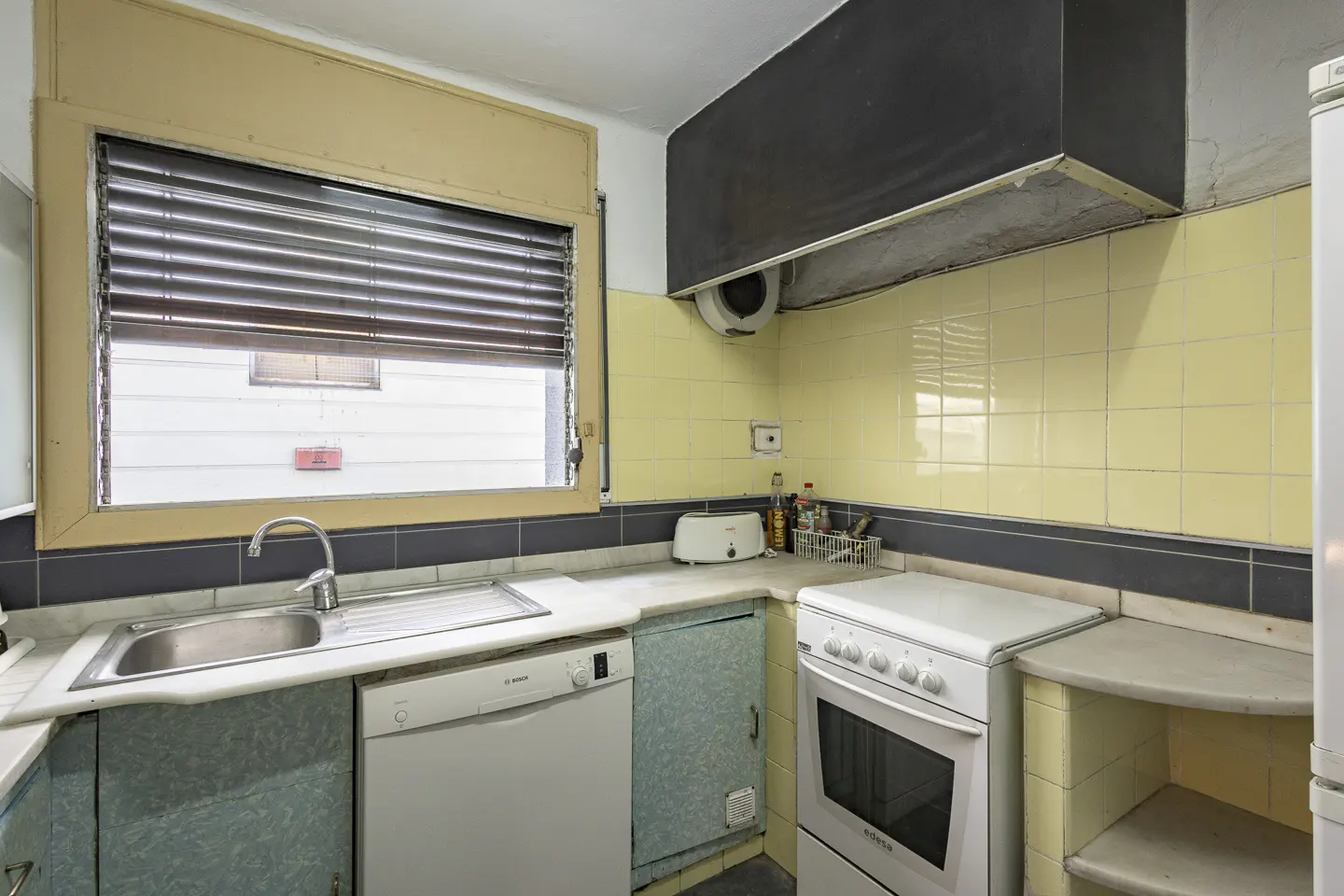 A kitchen with yellow tiled walls, a black range hood, a white stove, and a sink under a window with closed blinds.