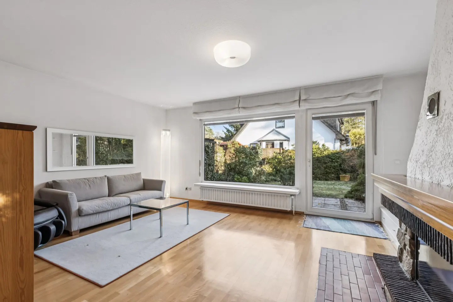 Bright living room with a gray sofa, glass table, and a large window overlooking a green backyard. A fireplace is visible on the right.