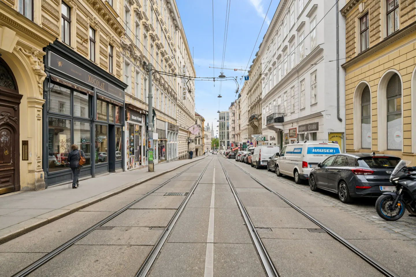 Street view in Vienna, Austria, with tram tracks running down the center. Buildings line the street, with shops and parked cars visible.