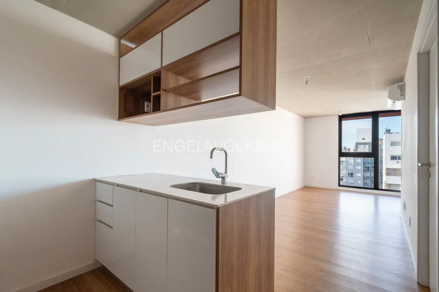 Modern apartment interior with white walls, wood floors, and a kitchen island with a sink and wood cabinets. A large window shows a city view.
