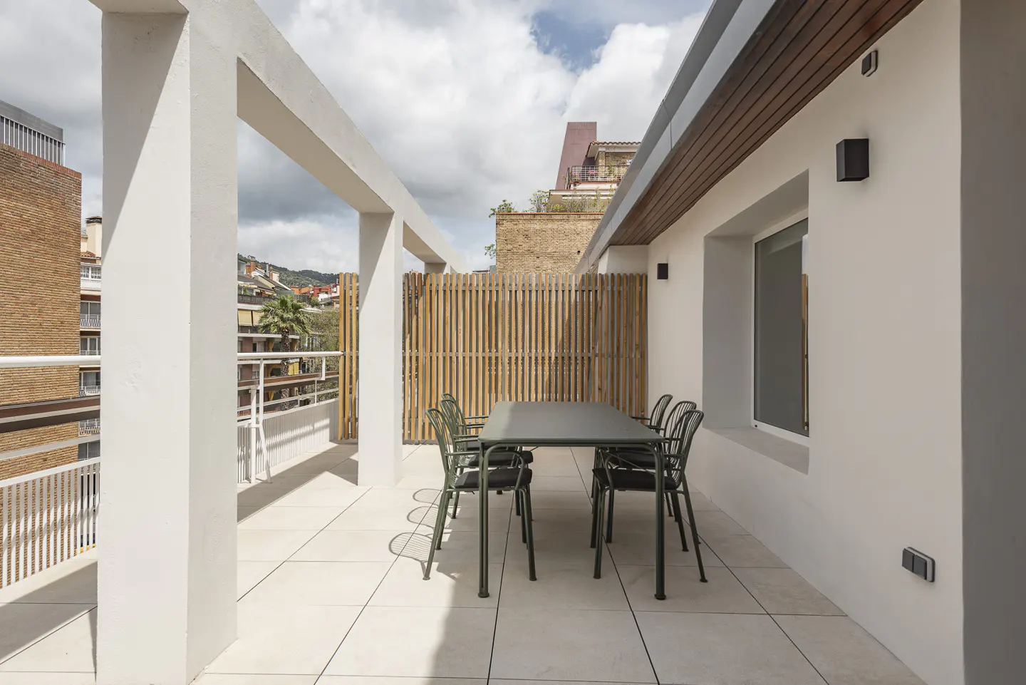Outdoor patio with a green table and chairs on a tiled floor. A bamboo fence and white pillars provide privacy and structure. City views in the background.