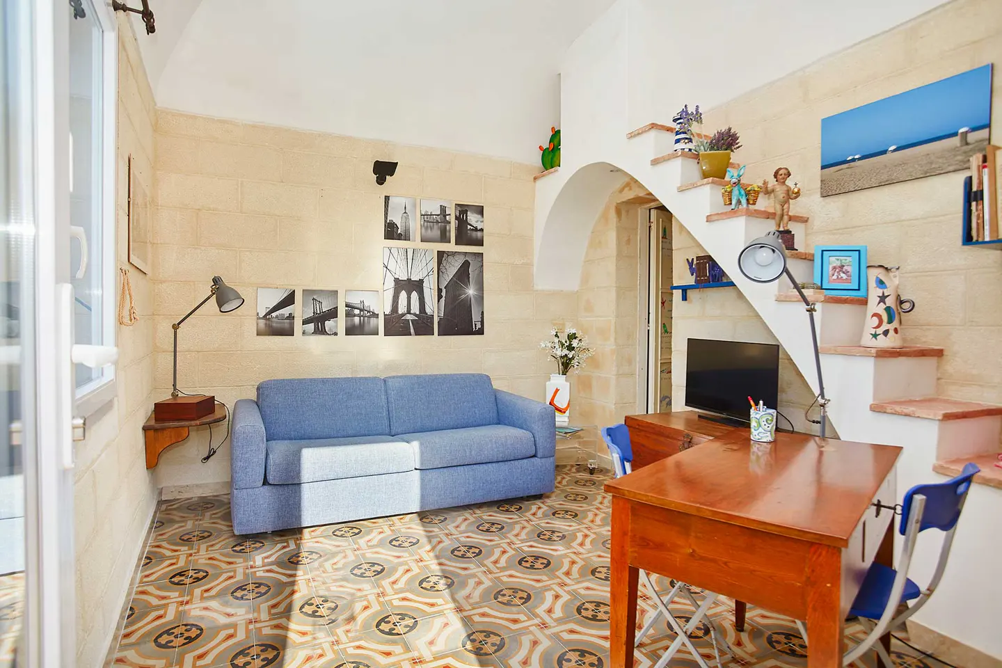 Living room with blue sofa, patterned tile floor, and desk. Stairs with decorations lead up. Black and white bridge photos on the wall.