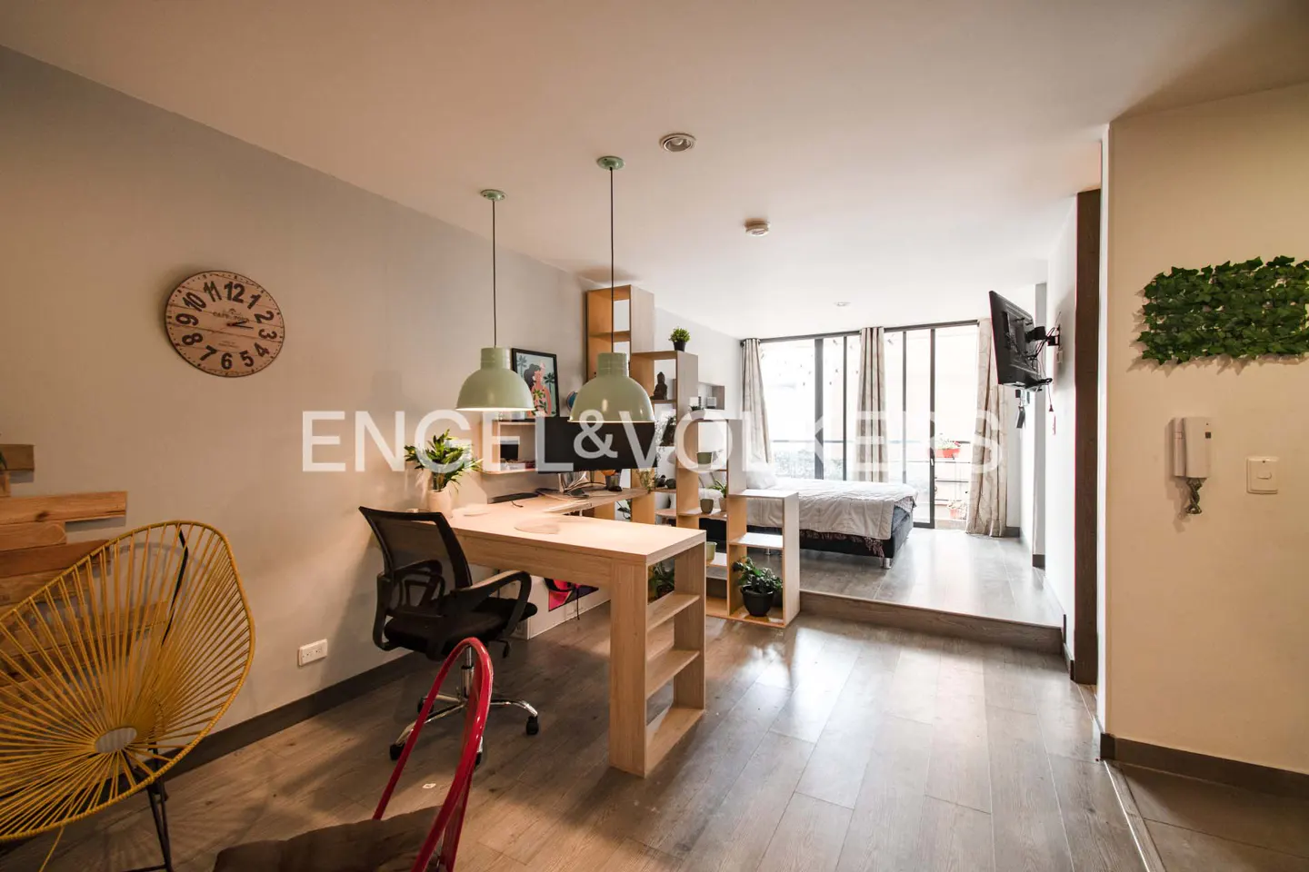 A modern studio apartment with a desk, bed, and balcony. The walls are light gray, and the floors are wood. There is a clock on the wall.