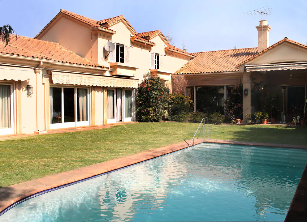 Backyard view of a two-story beige house with a red tile roof and a blue swimming pool in the foreground.