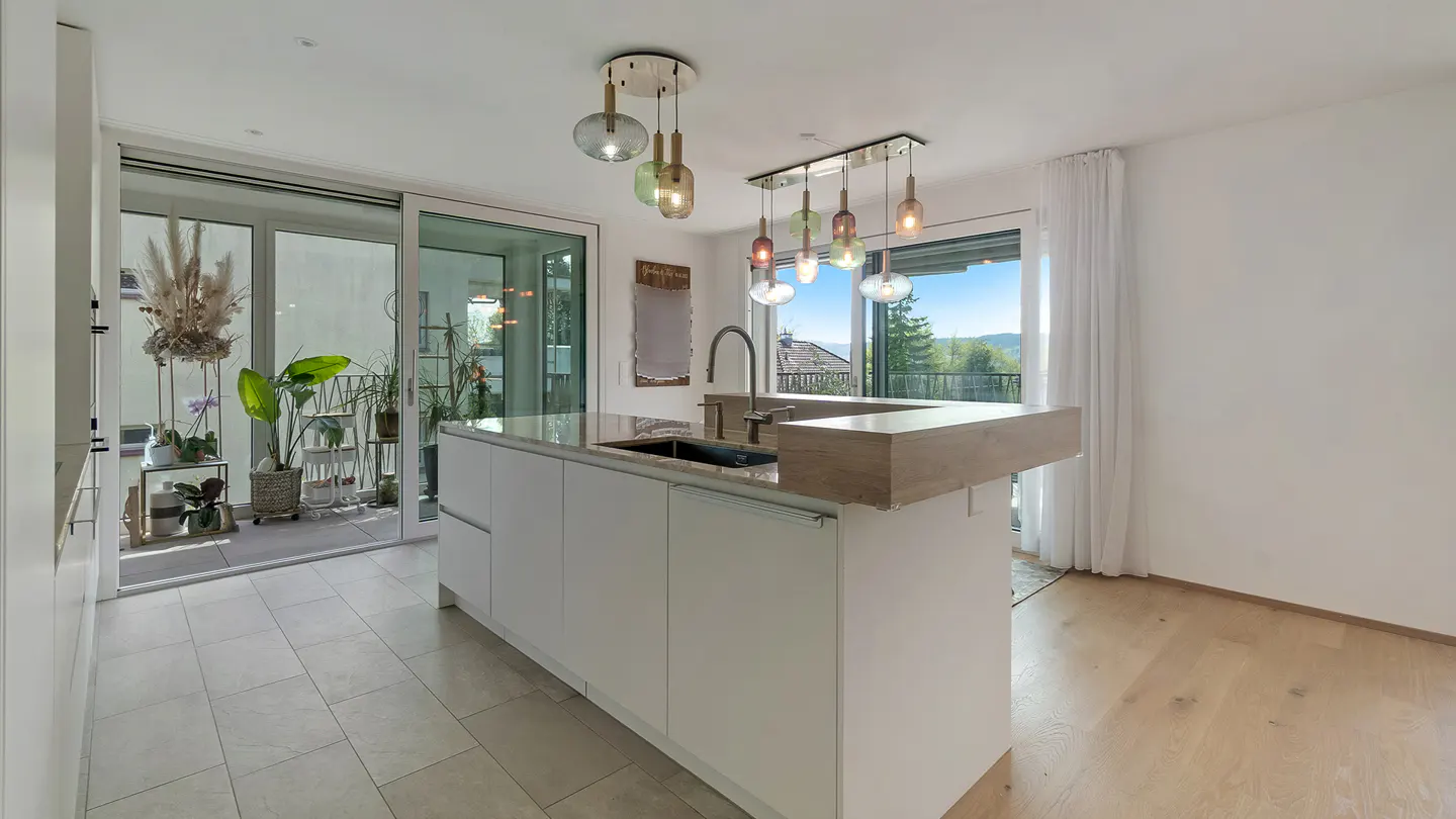 Bright kitchen with white cabinets, a stone countertop island, and pendant lighting. Sliding glass doors lead to a plant-filled balcony.
