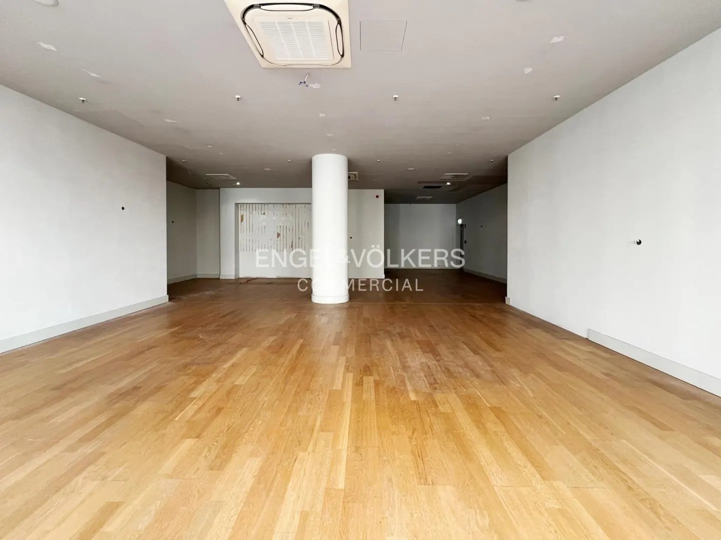 Empty commercial space with light wood floors, white walls, and a central support column. An air conditioning unit is visible on the ceiling.