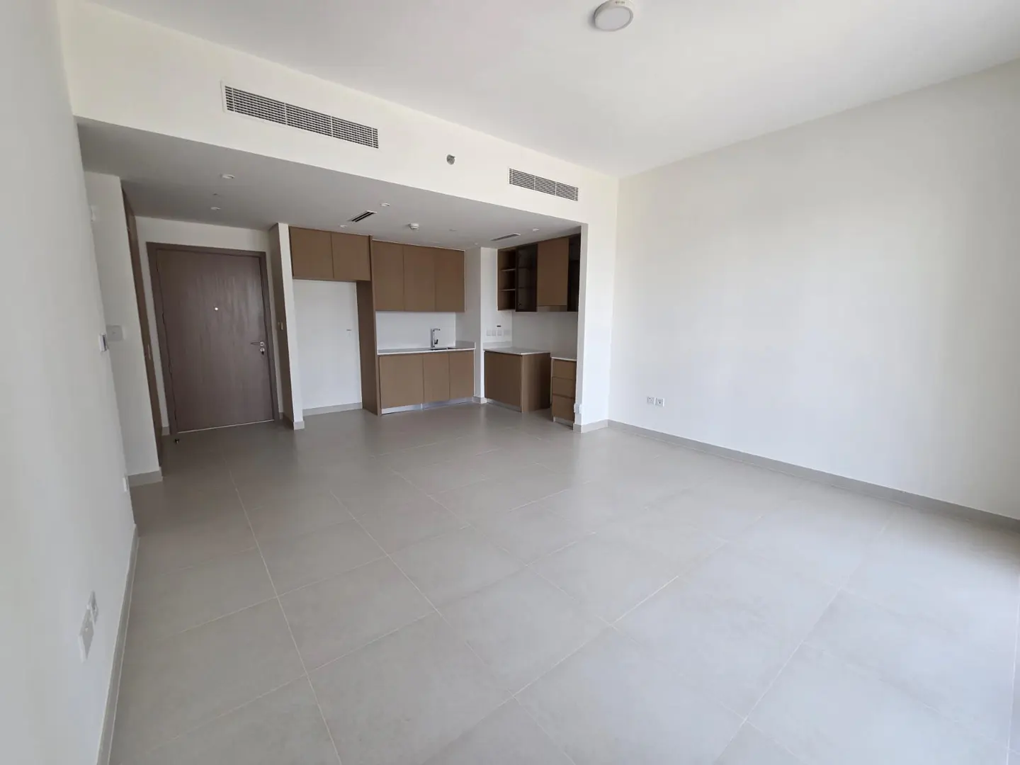 An empty, bright apartment with white walls, gray tile floors, and a brown kitchen area. A brown door is on the left.