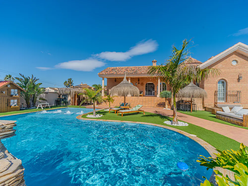 Exterior view of a luxury home with a blue swimming pool, palm trees, and lounge chairs under straw umbrellas.