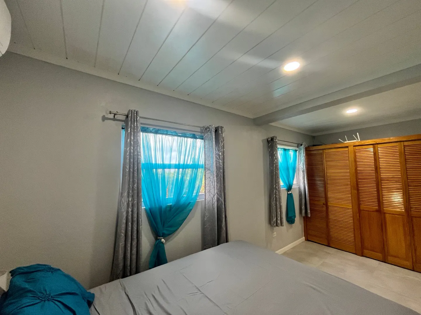 Bedroom with gray walls, a gray bedspread, and two windows with blue sheer curtains. A wooden closet is on the right.