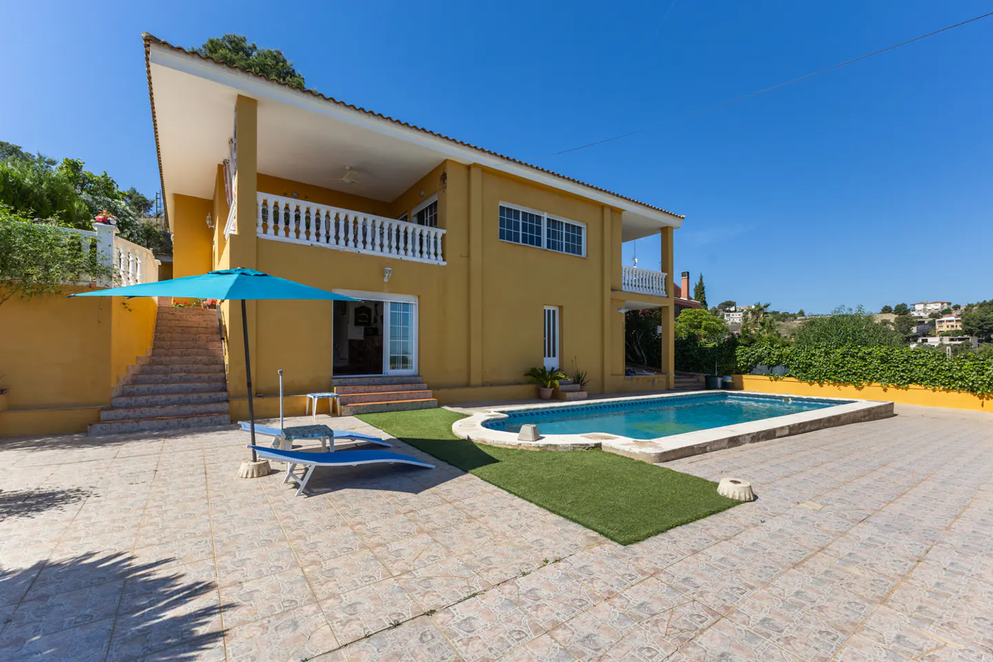 Two-story yellow house with a pool, blue umbrella, and lounge chairs on a tiled patio under a clear blue sky.