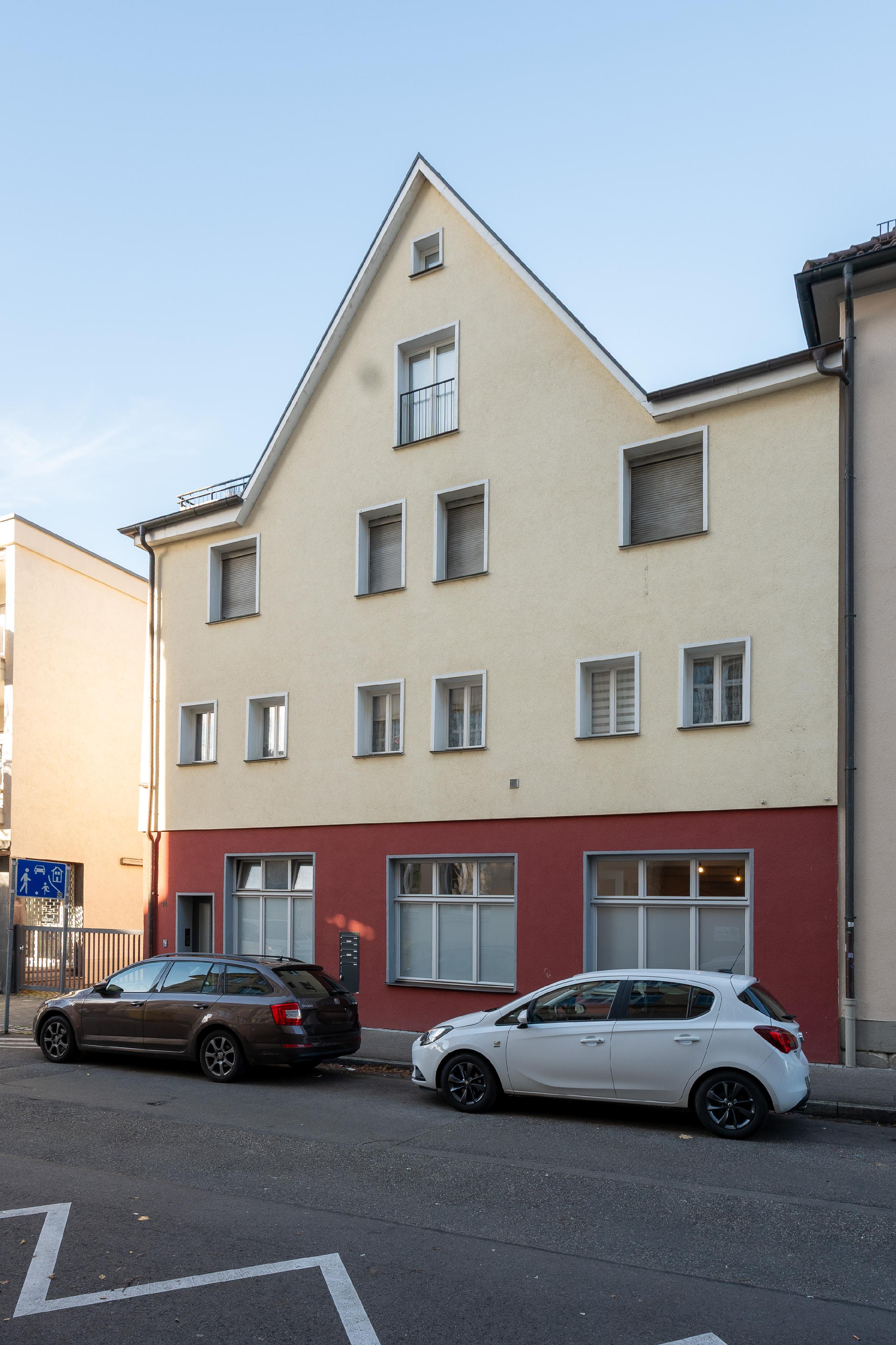 A three-story building with a cream upper facade and red ground floor, featuring multiple windows and parked cars in front. [AI-generated text]
